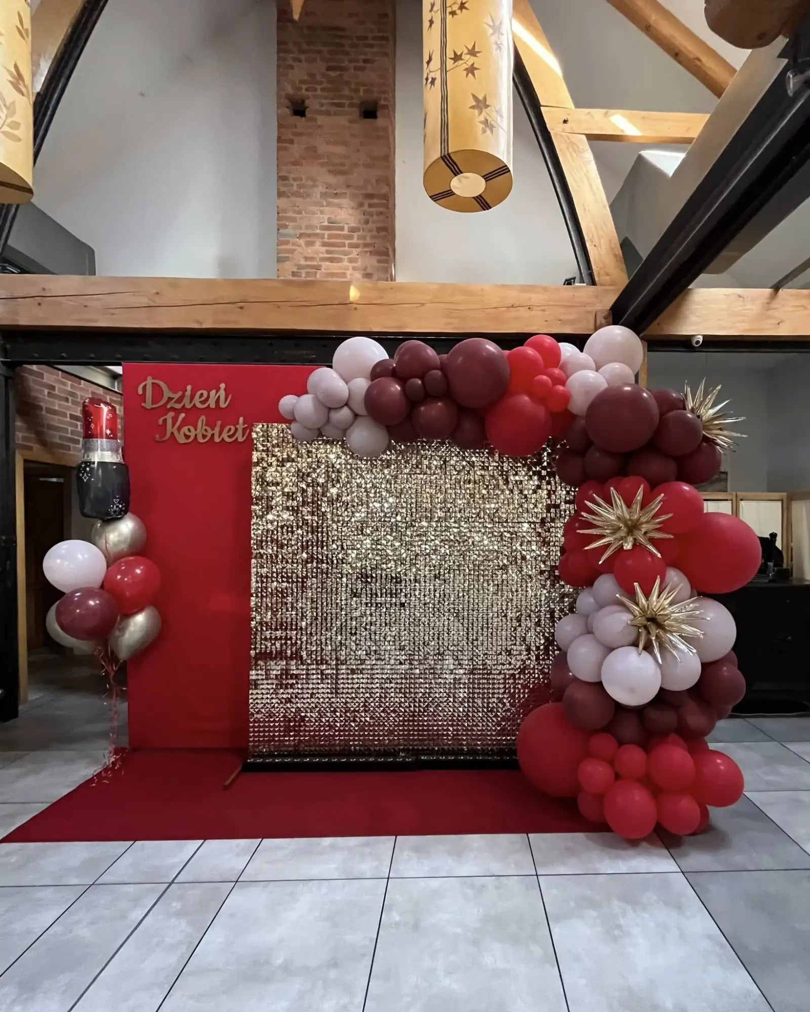 Błyszcząca złota tło z cekinami, obramowane łukiem czerwonych, bordowych i białych balonów z złotymi akcentami w kształcie rozbłysków gwiazd.