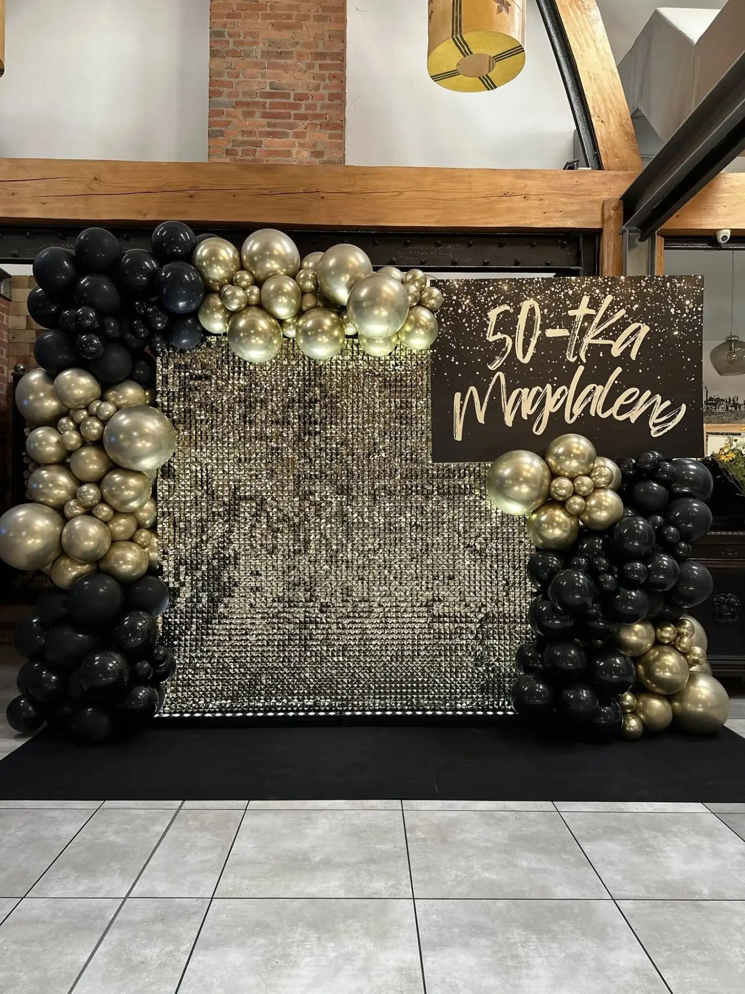 Błyszczący łuk balonowy w kolorach czarnym i złotym z migotliwym tłem, z grupami metalicznych balonów w różnych rozmiarach.