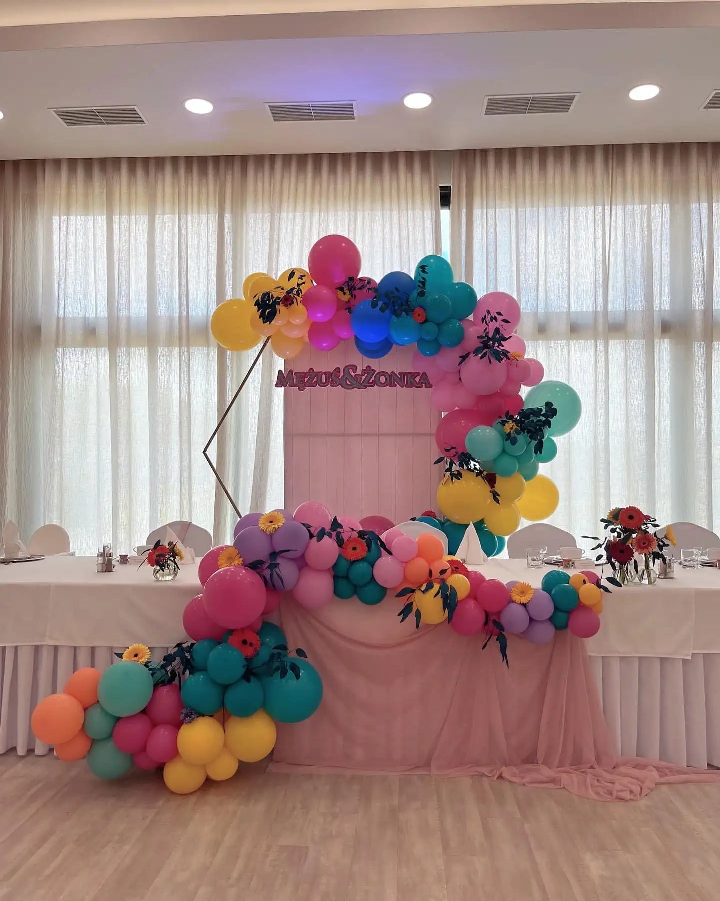 Wibrujący, wielobarwny łuk balonowy w odcieniach różu, żółtego, turkusu i fioletu z czarnymi kwiatowymi akcentami.